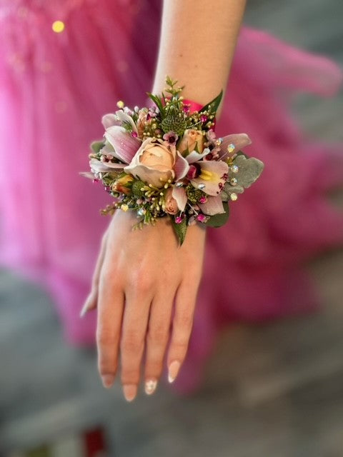 A floral wristlet corsage featuring large blush-spray rose buds centered among delicate pink cymbidium orchids. Accented with pink wax flower, scabiosa buds, seeded eucalyptus sprigs, white dusty miller leaves and shimmering rhinestone sprays, all tied with a fuchsia pink-colored satin ribbon.