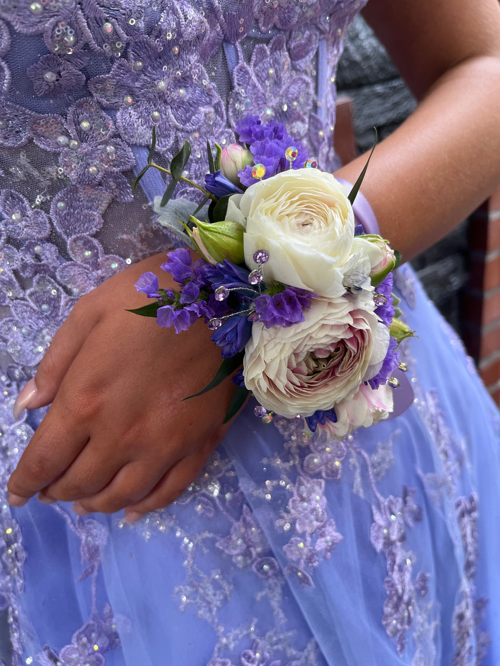 A floral wristlet corsage featuring a large blush-pink ranunculus centered among delicate pink spray rose buds. Accented with blue hyacinth blooms, purple statice, baby blue eucalyptus sprigs, and shimmering rhinestone sprays, all tied with a lilac-colored satin ribbon.