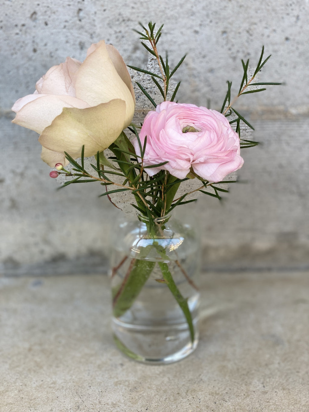 A close up of a clear glass bud vase with a blush rose and a blush ranunculus bloom and wax flower green foliage.