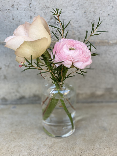 A close up of a clear glass bud vase with a blush rose and a blush ranunculus bloom and wax flower green foliage.