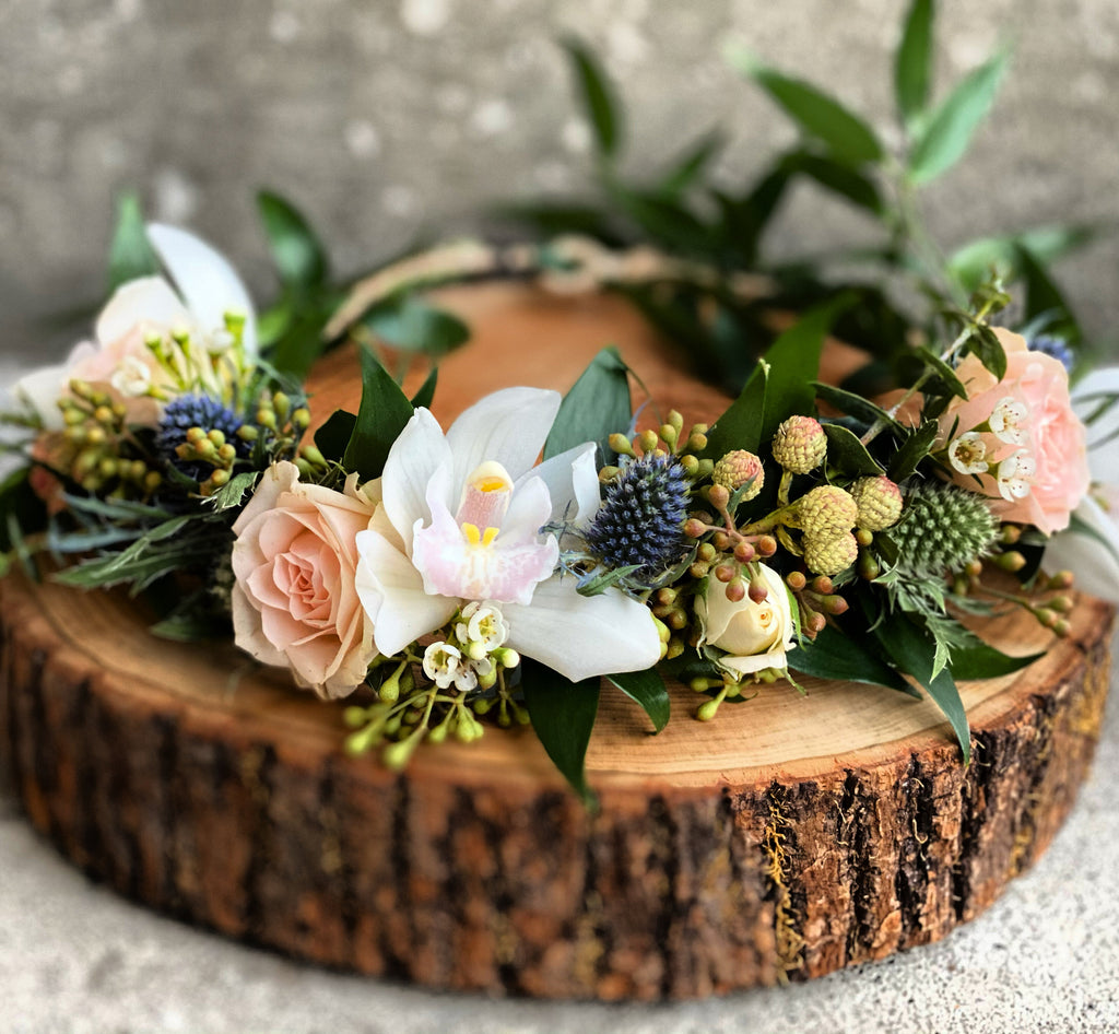 A close-up of a fresh floral crown featuring blush spray roses, blue eryngium, and white cymbidium orchids. Accented with brunia balls, seeded eucalyptus, and Italian ruscus, resting on a rustic wooden round slab.