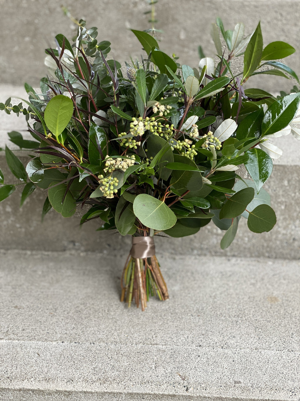 A close up of a textured bridesmaid attendant bouquet featuring a mixture dark green glossy leaves and seeded eucalyptus varieties.