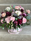 A lush mixed flower arrangement in a pink hat box. The design features soft blush Mondial roses and carnations contrasted with deep burgundy ranunculus. Spiky blue eryngium and hot pink stock add height, while delicate pink wax flowers, silver-toned eucalyptus, and dark agonis foliage provide rich texture.