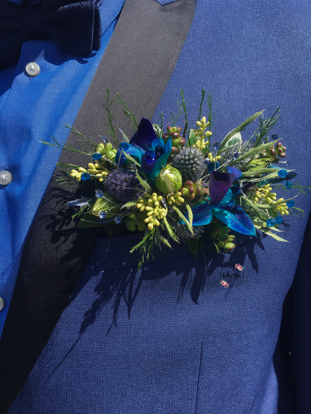 A floral boutonniere styled pocket square featuring vibrant blue dendrobium orchids, blue eryngium thistle, a pale rose bud, textured seeded eucalyptus, pops of blue rhinestone sprays, all bound with a navy blue hand-dyed silk ribbon.