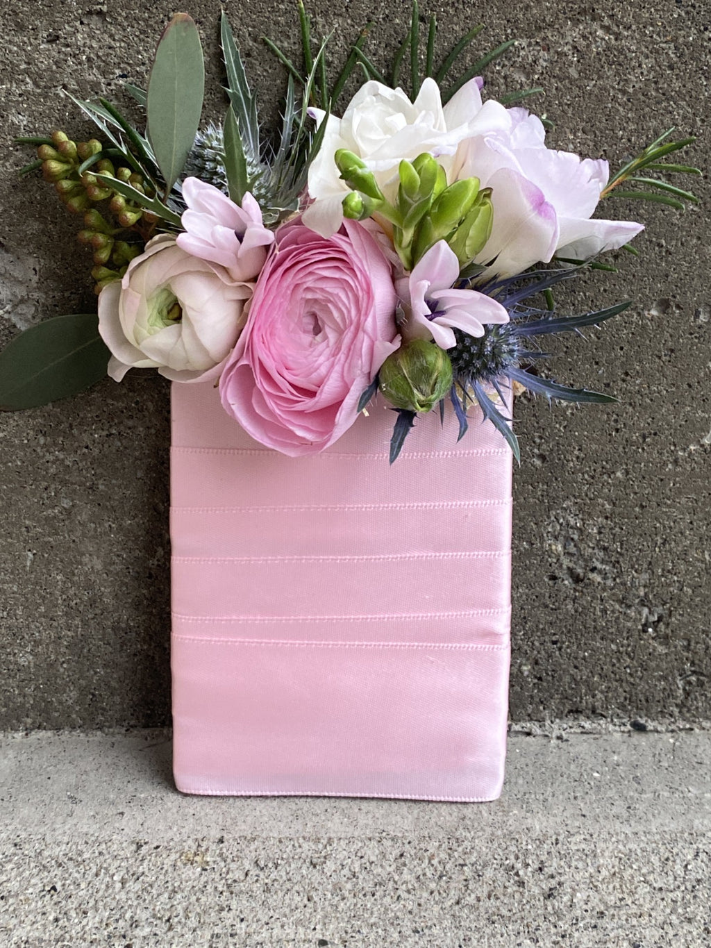 A floral boutonniere styled pocket square featuring pale pink ranuculus,  lavender tipped freesia and buds, pink hyacinth blooms, blue eryngium, and textured seeded eucalyptus, all bound with a light pink satin ribbon.