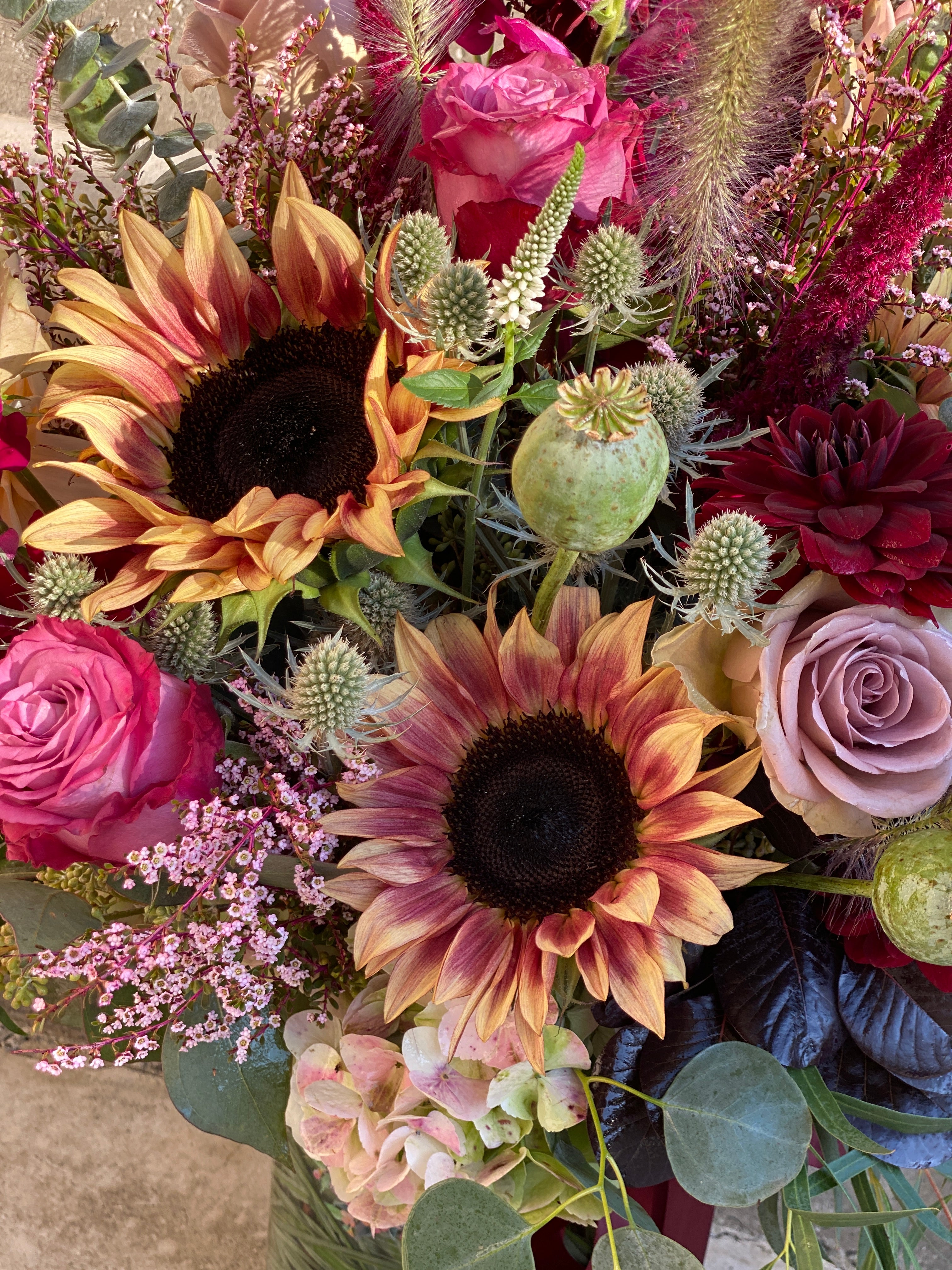 A close-up of a vibrant floral arrangement featuring two large sunflowers with orange and dark brown centers, pink roses, burgundy dahlias, light pink hydrangeas, and green foliage.