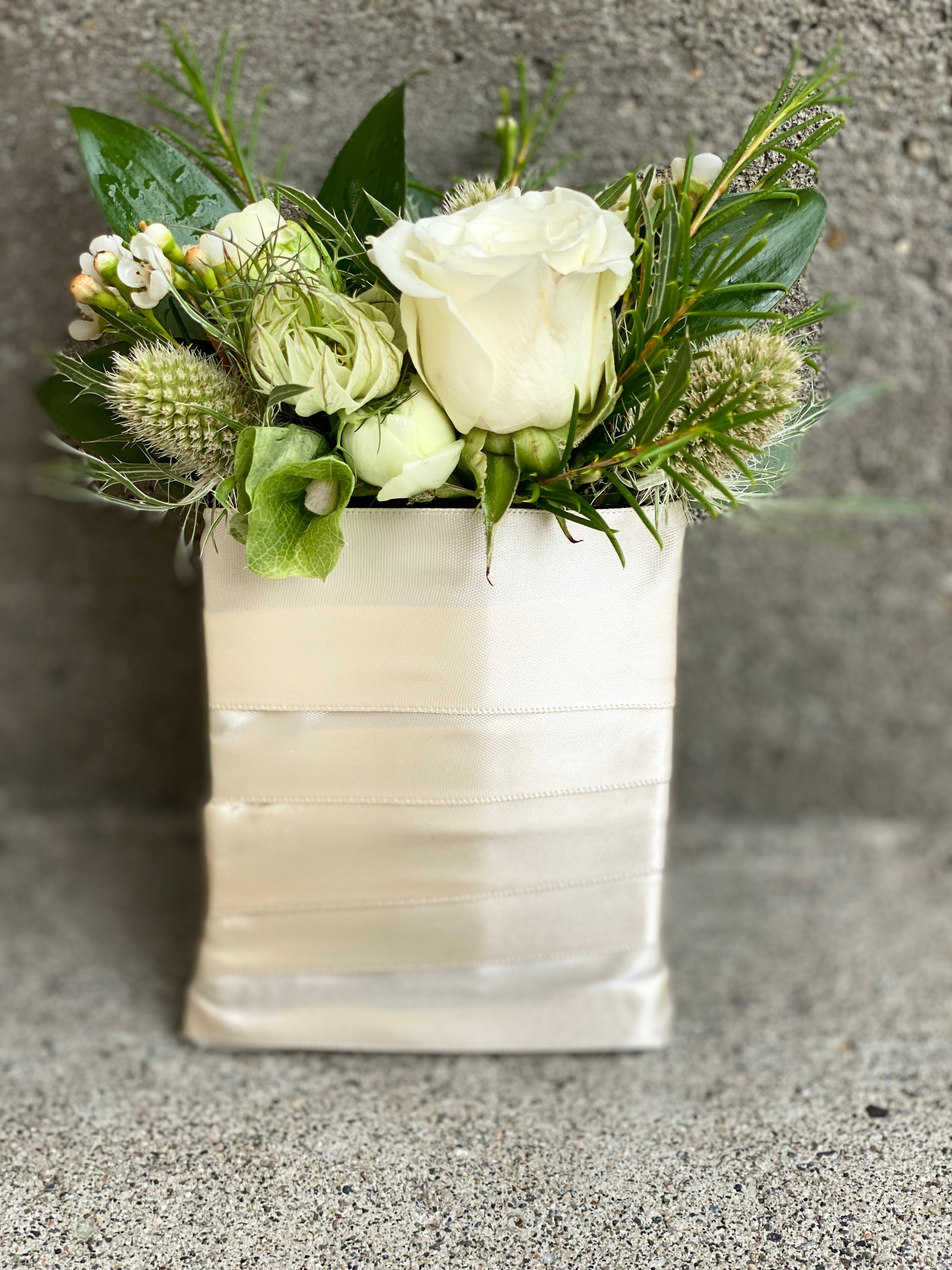 A floral boutonniere styled pocket square featuring a white spray rose,  nigella, ranunculus buds, and green lagoon eryngium tips, and dark green Italian Ruscus leaves, all bound with a cream satin ribbon around the base.