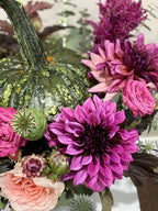 A close-up of a fall floral arrangement featuring a green speckled pumpkin, bright pink and magenta dahlias, blush pink roses, and green foliage.