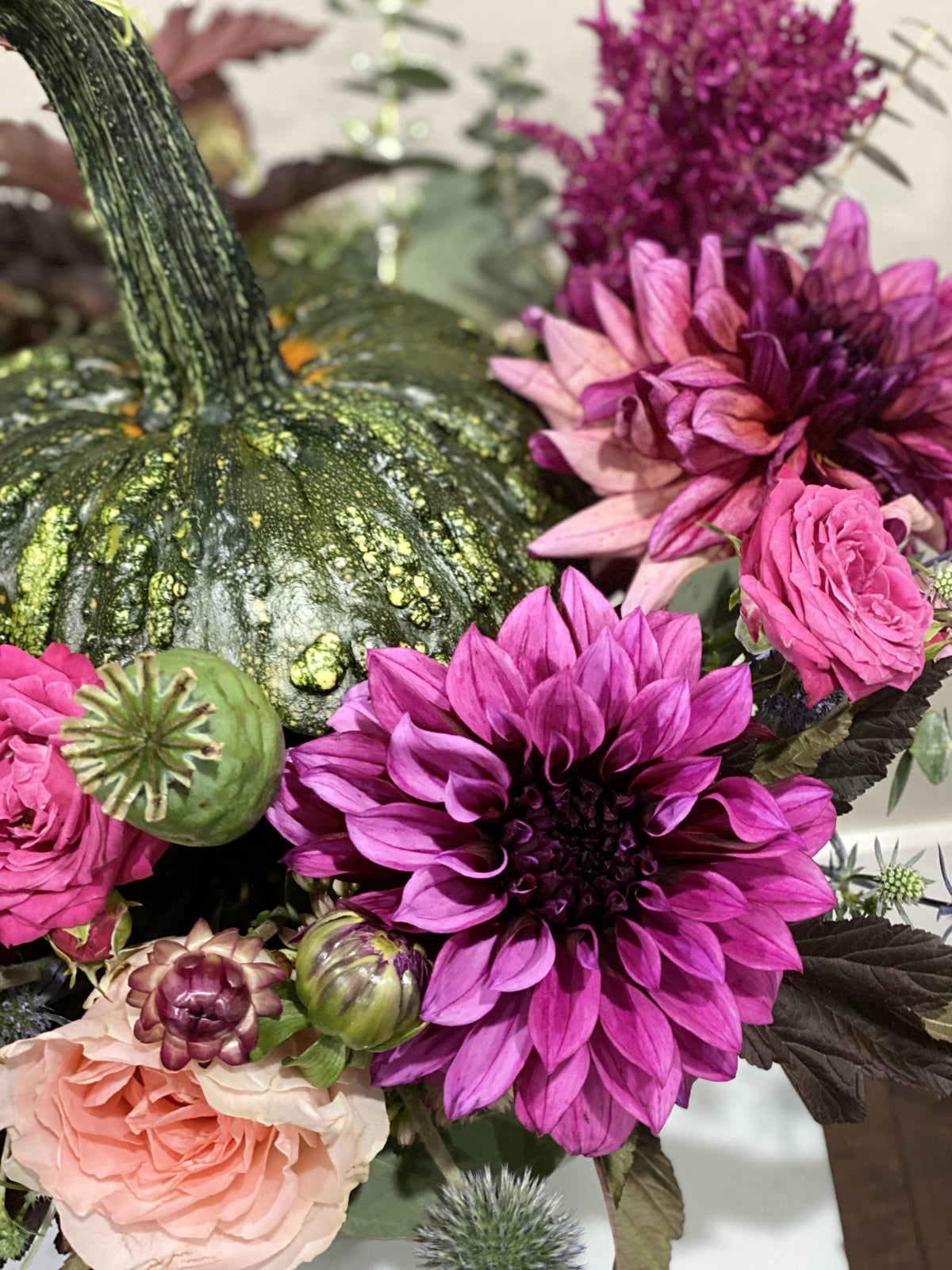 A close-up of a fall floral arrangement featuring a green speckled pumpkin, bright pink and magenta dahlias, blush pink roses, and green foliage.