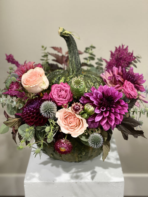 A decorative floral arrangement featuring a small green pumpkin nestled among pink and purple flowers, including roses, dahlias, and thistle-like blooms, displayed on a white pedestal against a neutral indoor background.
