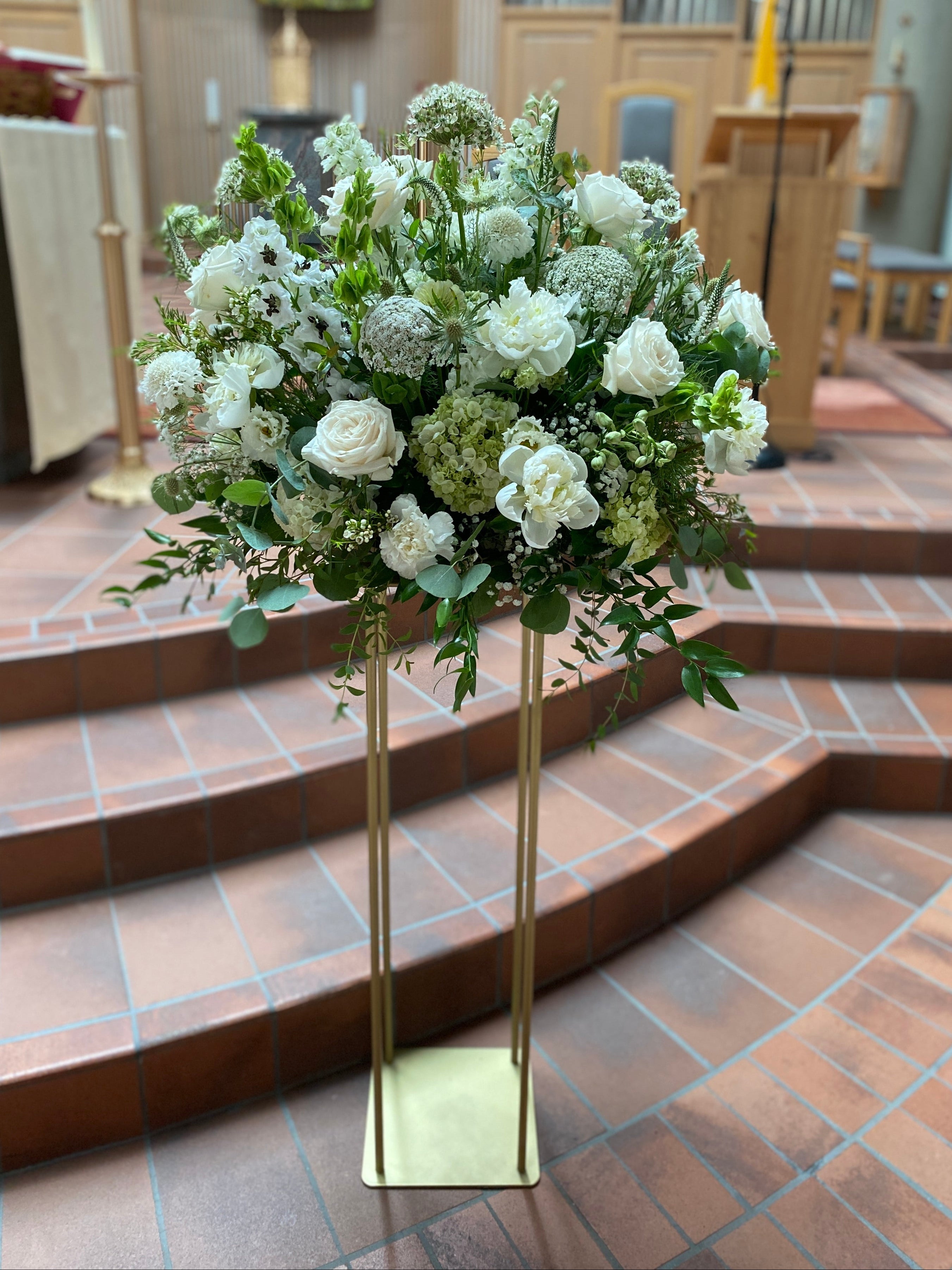 A tall floral arrangement of white flowers and green foliage displayed on a slim gold metal stand in a church interior with brick steps and wooden pews in the background.