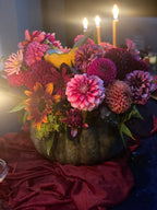 A large decorative pumpkin filled with a dense floral arrangement of pink, magenta, orange, and burgundy flowers, styled on draped burgundy fabric with lit candles in the background.