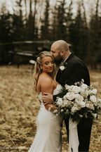 A bride and groom sharing an intimate moment in a vast, open field during their elopement. The bride holds a large, lush white bouquet featuring white roses, ranunculus, delicate bridal spirea, and various textured green eucalyptus leaves