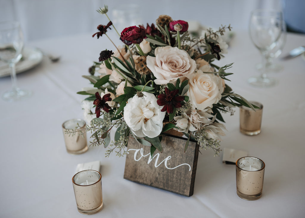 A lush wedding centerpiece in a low, gold footed vase featuring dusty-pink Quicksand roses, deep burgundy ranunculus, and dark chocolate cosmos. The arrangement is accented by blush bridal protea, textured dried scabiosa pods, and trailing eucalyptus and ruscus greenery.