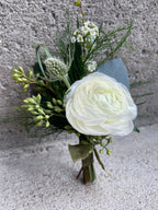 A formal boutonniere featuring a focal white ranunculus bloom accented by spiky green eryngium and delicate white bridal spirea. The boutonniere is framed by wispy loops of green bear grass, textured tree fern, and small seeded eucalyptus tips.
