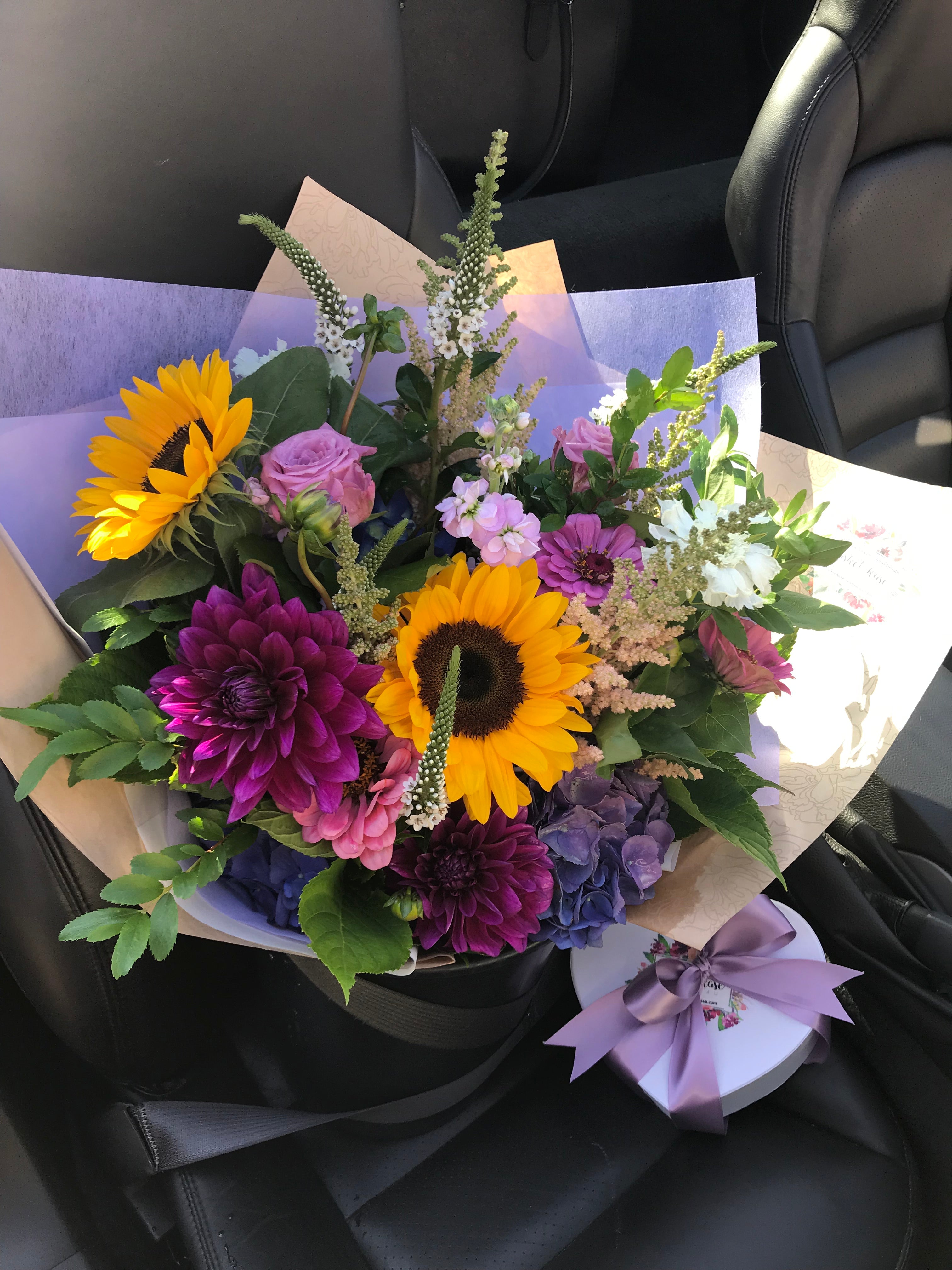 Bouquet of Summer flowers with Sunflowers, pink Roses, and purple Dahlia blooms wrapped in kraft paper and tissue.