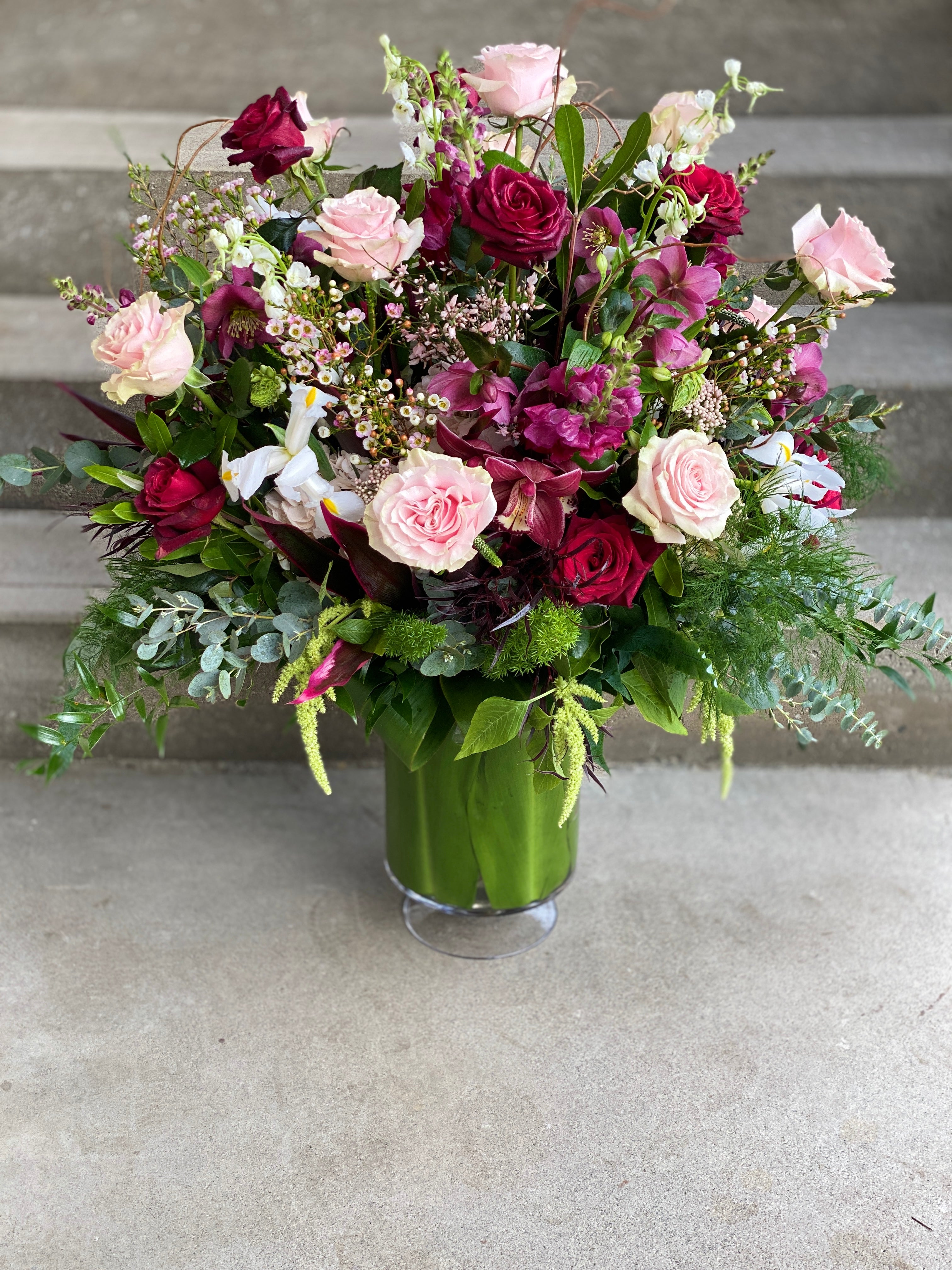 Large statement arrangement in a footed clear vase, featuring blush and red garden rose, burgundy cymbidium orchids, red hellebores, pink wax flower, white iris, white larkspur, and burgundy snap dragons, accented with lime green amaranthus, burgundy and green Ti  leaves, plumosa and tree fern, and eucalyptus greenery.