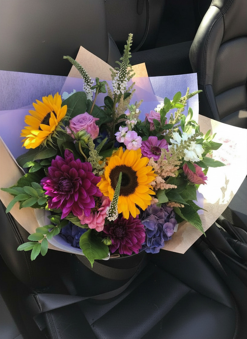 A vibrant mixed flower bouquet featuring two yellow sunflowers, deep purple dahlias, pink roses, and purple hydrangeas, wrapped in kraft paper and placed on a black car seat.
