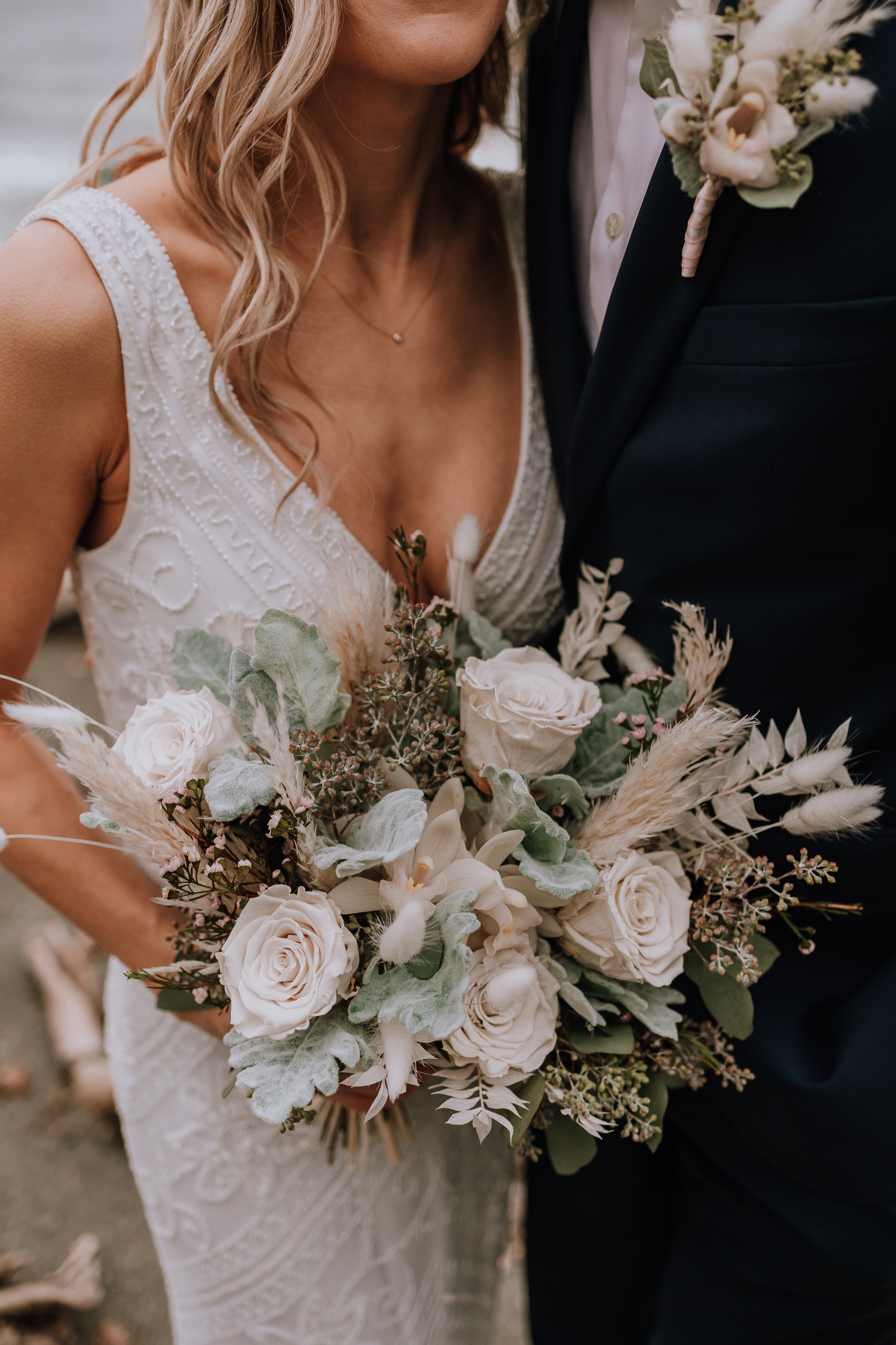 A close-up of a bride holding her bouquet of white preserved roses, orchids, dusty miller, and bunny tails, and wax flowers and eucalyptus varieties while she stands beside the groom.