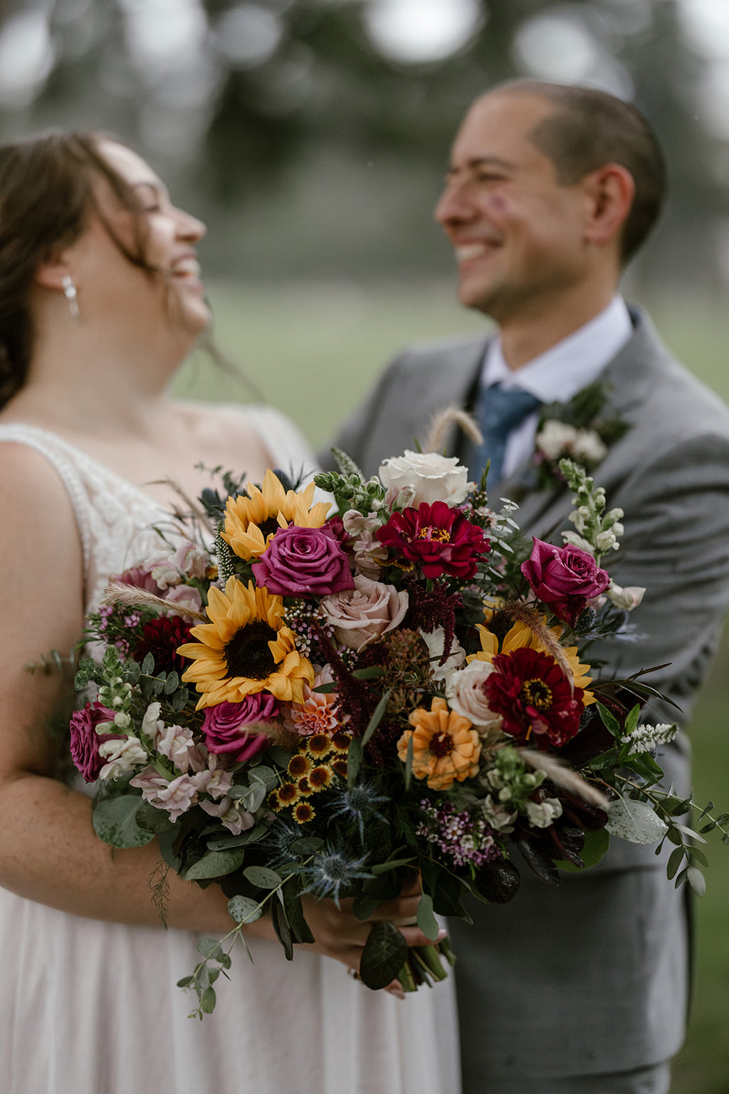 Close-up of a laughing bride and groom. The bride carries a large, colorful bridal bouquet featuring bright yellow sunflowers, pink zinnias, fuchsia roses, and various silver-green eucalyptus leaves."