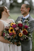 Close-up of a laughing bride and groom. The bride carries a large, colorful bridal bouquet featuring bright yellow sunflowers, pink zinnias, fuchsia roses, and various silver-green eucalyptus leaves."