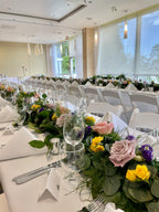 Decorated event hall with tables set for a gathering, featuring blush roses, yellow spray roses, purple lisianthus and eucalyptus greenery garland down the center of the long tables and white chairs.