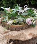 A close-up of a fresh floral crown featuring blush bridal protea, green eryngium, and white wax flower. Accented with seeded eucalyptus, and Italian ruscus, resting on a rustic wooden round slab on burlap.