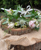 A close-up of a fresh floral crown featuring blush bridal protea, green eryngium, and white wax flower. Accented with seeded eucalyptus, and Italian ruscus, resting on a rustic wooden round slab on burlap.