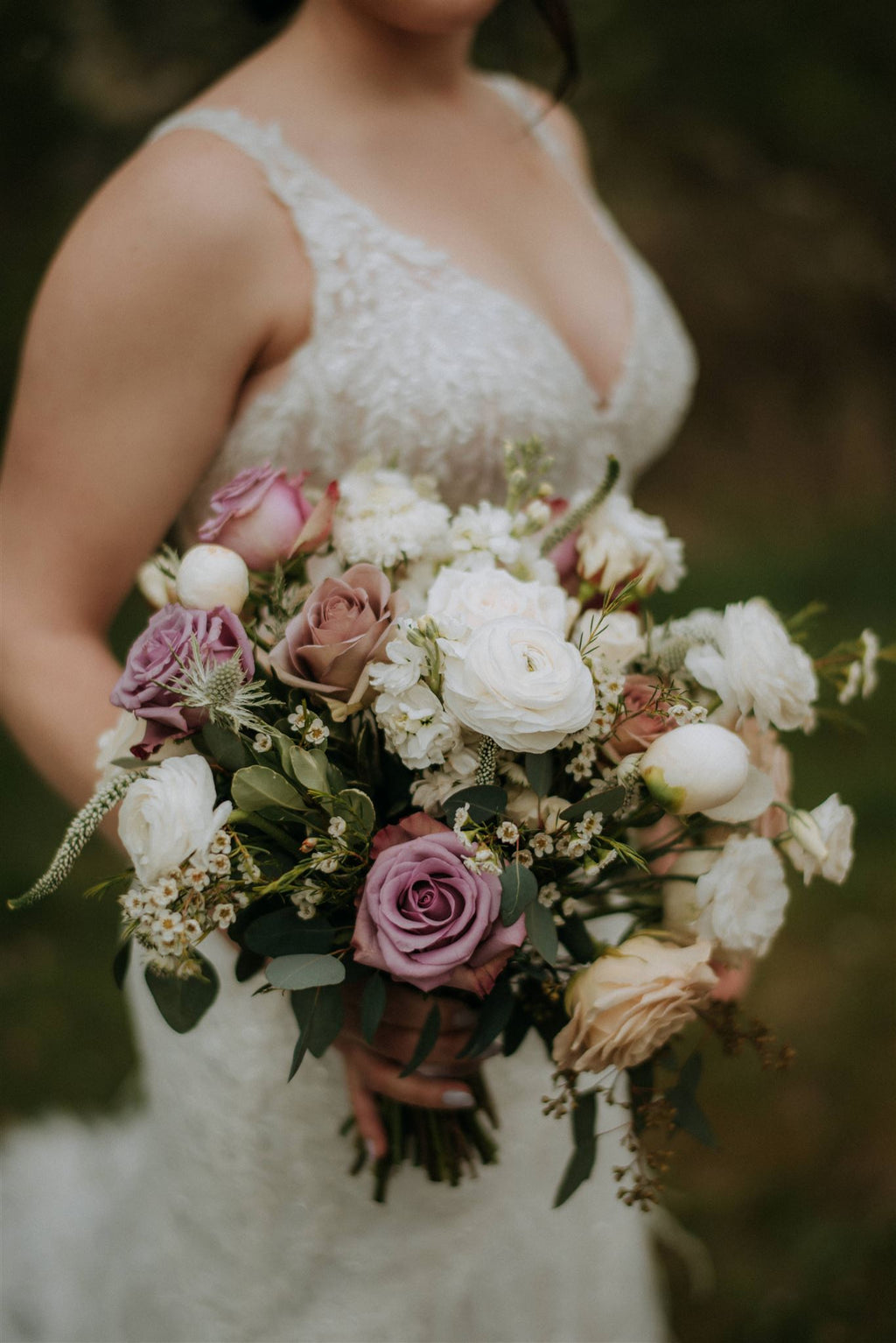 Close-up of a bride in a sunlit garden holding her lush bouquet. The bouquet features a blend of crisp white ranunculus, Westminster garden roses, and muted Amnesia mauve roses paired with soft blush blooms and greenery.