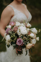 Close-up of a bride in a sunlit garden holding her lush bouquet. The bouquet features a blend of crisp white ranunculus, Westminster garden roses, and muted Amnesia mauve roses paired with soft blush blooms and greenery.