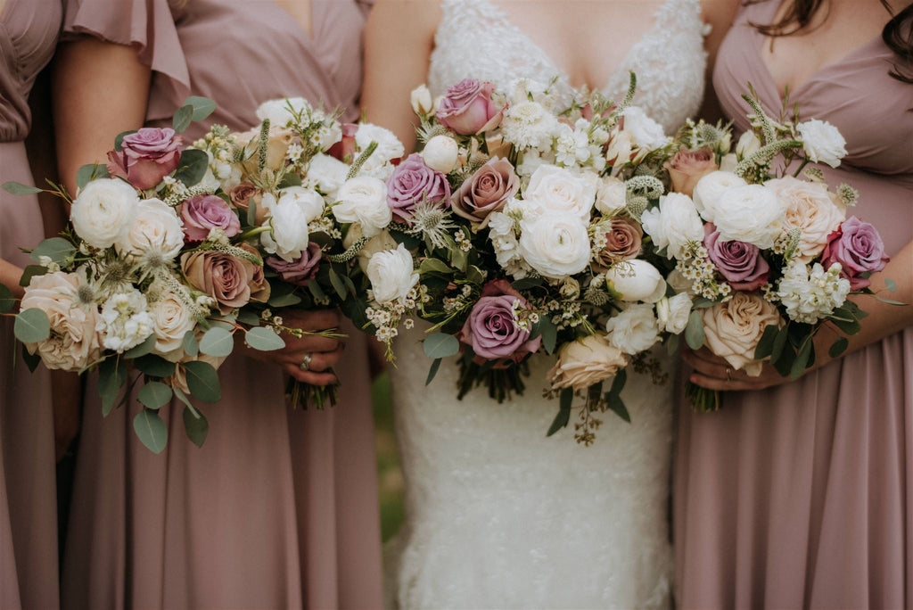Close-up of a bride and bridesmaids in a sunlit garden holding lush bouquets. The arrangements feature a blend of crisp white ranunculus, Westminster garden roses, and muted Amnesia mauve roses paired with soft blush blooms and greenery.