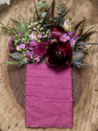 A moody and textured floral boutonniere-style pocket square featuring a deep burgundy ranunculus, blue eryngium, and a mauve mum. Accented with delicate wax flowers, glossy camellia leaves, and eucalyptus, all bound together with a mulberry-colored, hand-dyed silk ribbon.