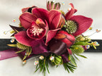A floral wrist corsage on a black slap band featuring burgundy cymbidium orchids and peony buds. Accents include green eryngium, red astrantia, scabiosa buds, hebe tips, seeded eucalyptus, finished with burgundy organza and satin ribbon loops and shimmering silver rhinestone sprays