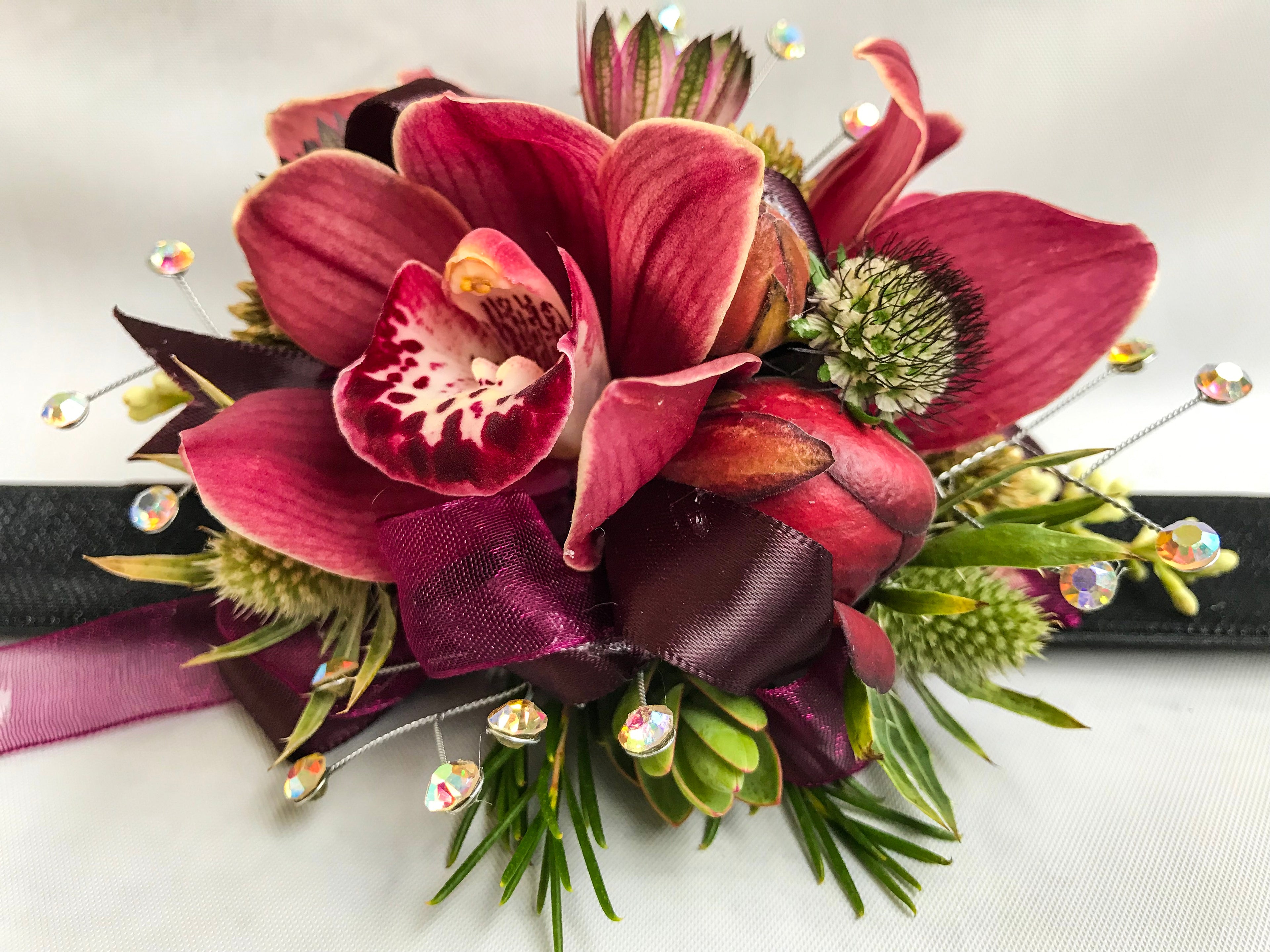 A floral wrist corsage on a black slap band featuring burgundy cymbidium orchids and peony buds. Accents include green eryngium, red astrantia, scabiosa buds, hebe tips, seeded eucalyptus, finished with burgundy organza and satin ribbon loops and shimmering silver rhinestone sprays