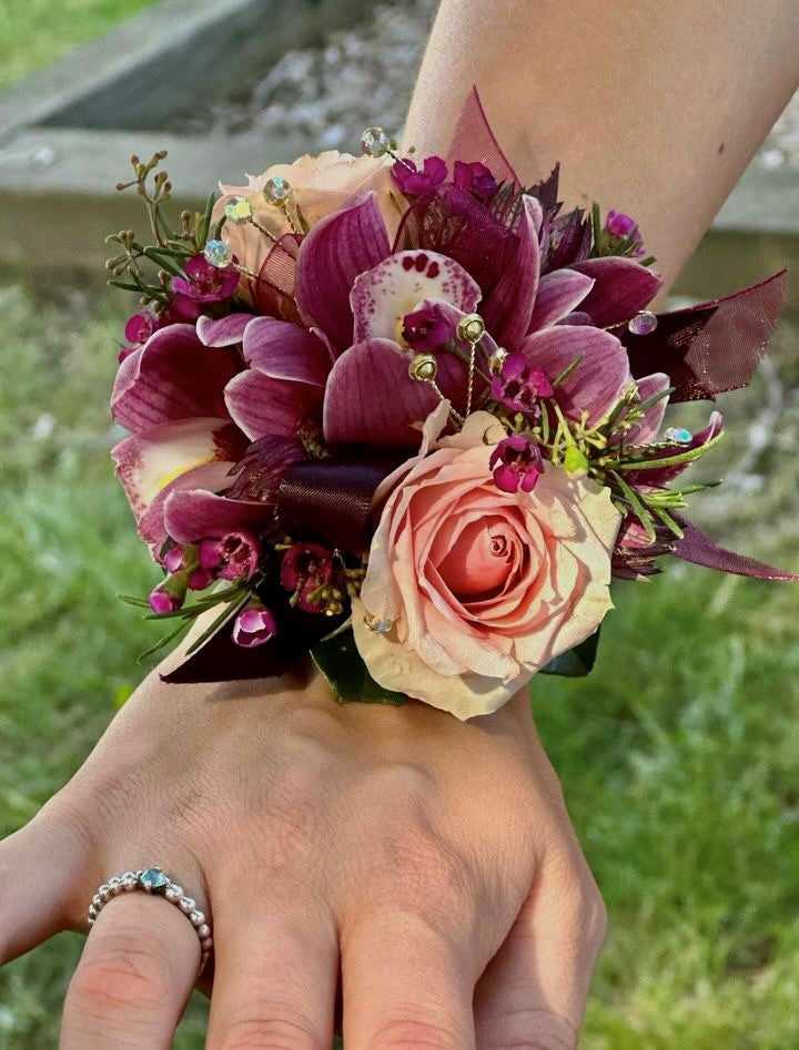 A floral wrist corsage on a black slap band featuring burgundy cymbidium orchids centered among peach spray roses and wax flower. Accents include burgundy astrantia tips, and seeded eucalyptus, finished with burgundy satin ribbon loops and shimmering rhinestone sprays.