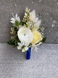 A formal boutonniere featuring a focal white ranunculus bloom and pale yellow spray roses accented by spiky green eryngium and delicate dried bunny tails and bleached italian ruscus. The boutonniere is finished with navy blue satin ribbon on the stems.