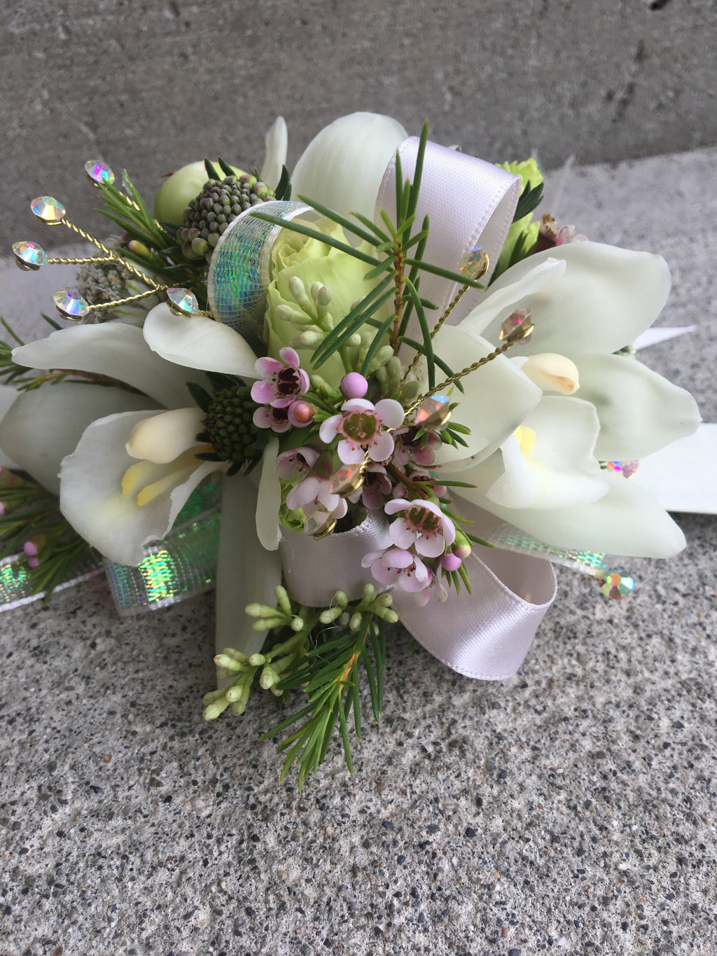 A floral wrist corsage on a white slap band featuring white cymbidium orchids centered among cream spray roses and peony buds. Accents include pink wax flower, and seeded eucalyptus, finished with blush and iridescent  ribbon loops and shimmering rhinestone sprays.