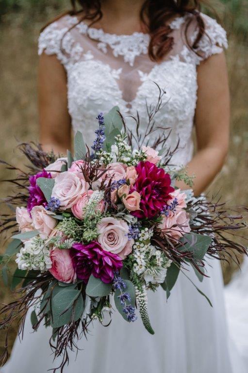 A bride in an intricate white lace wedding dress holds a lush bouquet of blush and fuchsia roses and dahlias, accented with silver dollar and seeded eucalyptus