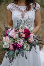 A bride in an intricate white lace wedding dress holds a lush bouquet of blush and fuchsia roses and dahlias, accented with silver dollar and seeded eucalyptus