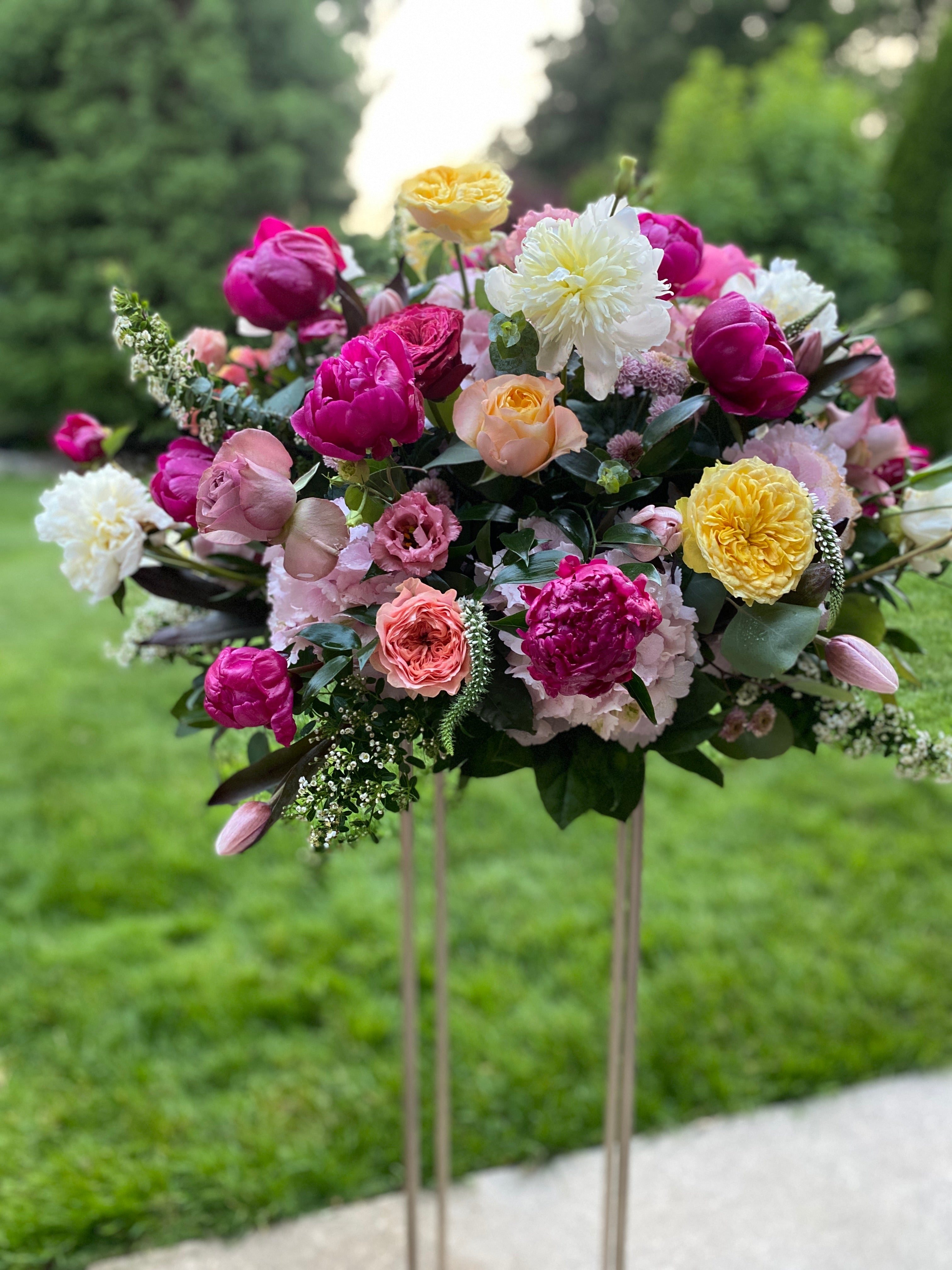 A large vibrant elevated flower arrangement nestled on top of a metal stand. Featuring fuchsia and white peonies, yellow garden roses, mauve roses and assorted greenery.