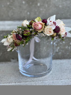 A close-up of a fresh floral crown dislayed on a glass vase featuring blush spray roses, white cymbidium orchids, peony buds, red astrantia, and white wax flower, tied with blush ribbon.