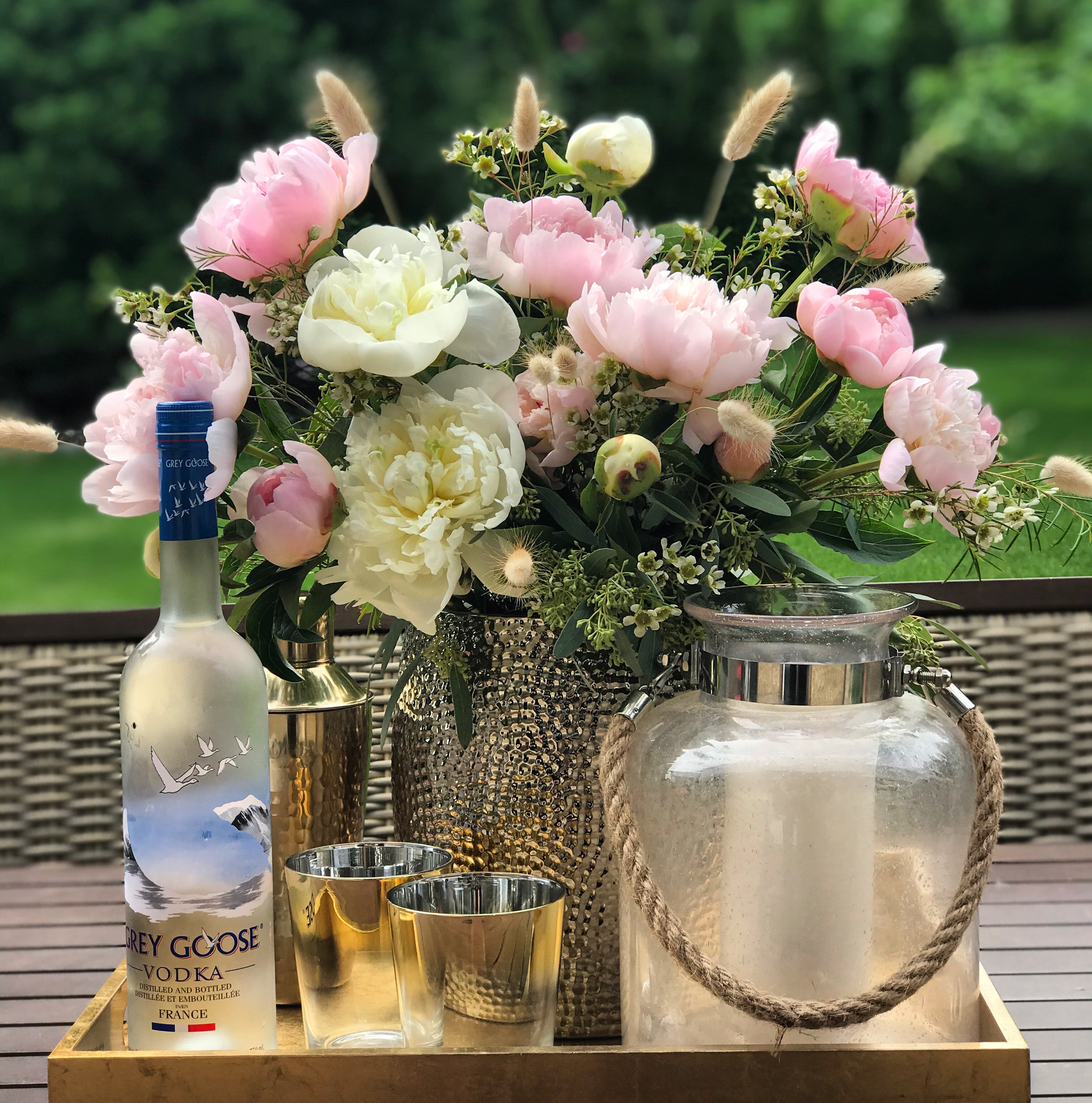 A large hammered metal vase designed with white and blush peonies, bunny tails, seeded eucalyptus and wax flower placed on a table with a grey goose vodka bottle, gold rimmed glasses, and a metal shaker.