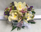 A floral wrist corsage on a white slap band featuring pale cream cymbidium orchids. Accents include pink wax flower, scabiosa buds, purple lavender tips, seeded eucalyptus, glossy green italian ruscus, finished with mauve and iridescent organza ribbon loops and shimmering lavender rhinestone sprays.
