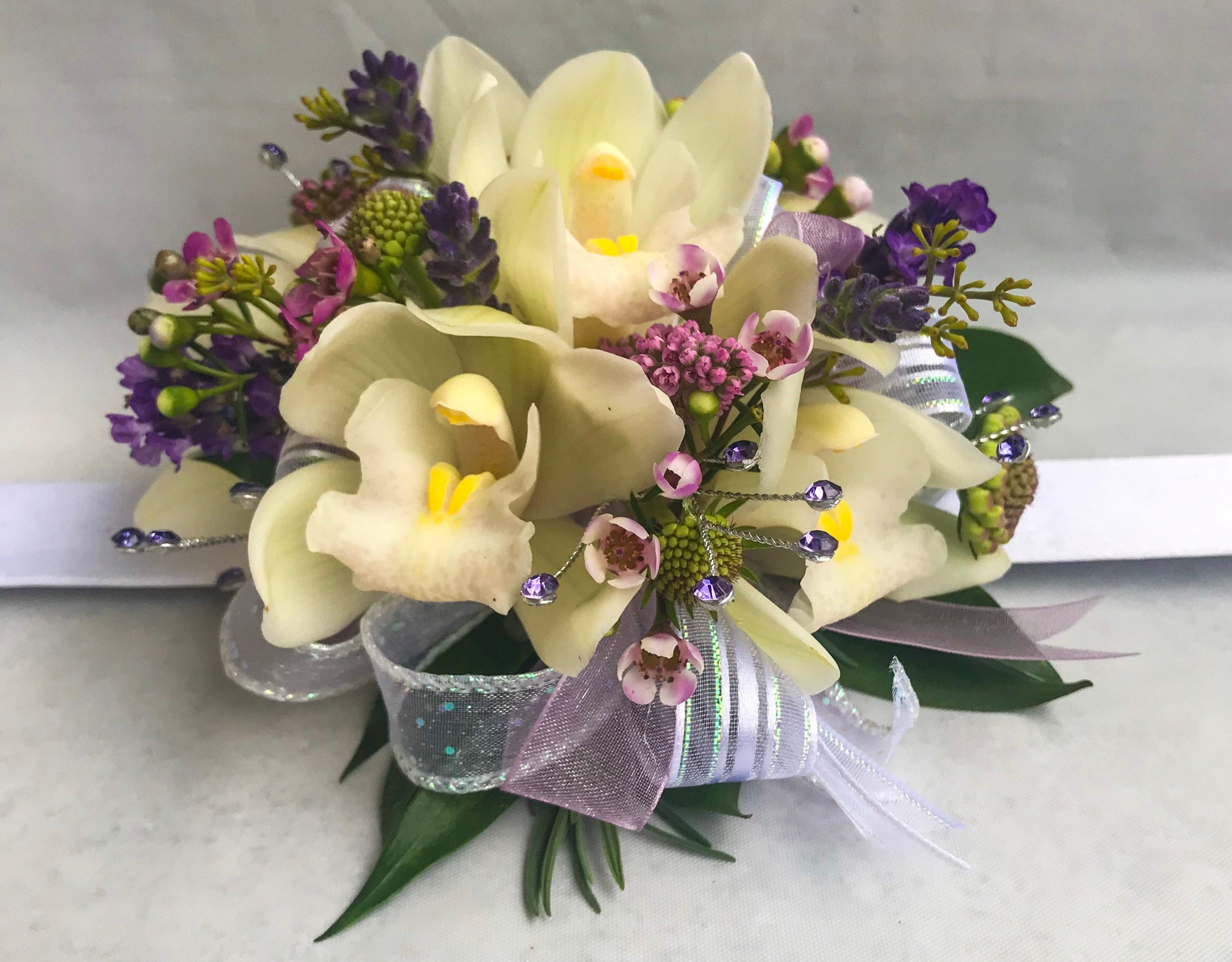 A floral wrist corsage on a white slap band featuring pale cream cymbidium orchids. Accents include pink wax flower, scabiosa buds, purple lavender tips, seeded eucalyptus, glossy green italian ruscus, finished with mauve and iridescent organza ribbon loops and shimmering lavender rhinestone sprays.