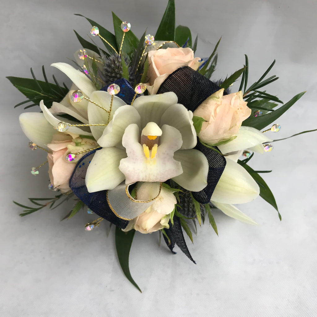 A floral wrist corsage on a cream slap band featuring pale cream cymbidium orchids centered among blush spray rose buds. Accents include glossy green italian ruscus and wax flower tips, blue eryngium, finished with cream and navy blue organza ribbon loops and shimmering rhinestone sprays.