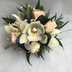 A floral wrist corsage on a cream slap band featuring pale cream cymbidium orchids centered among blush spray rose buds. Accents include glossy green italian ruscus and wax flower tips, blue eryngium, finished with cream and navy blue organza ribbon loops and shimmering rhinestone sprays.