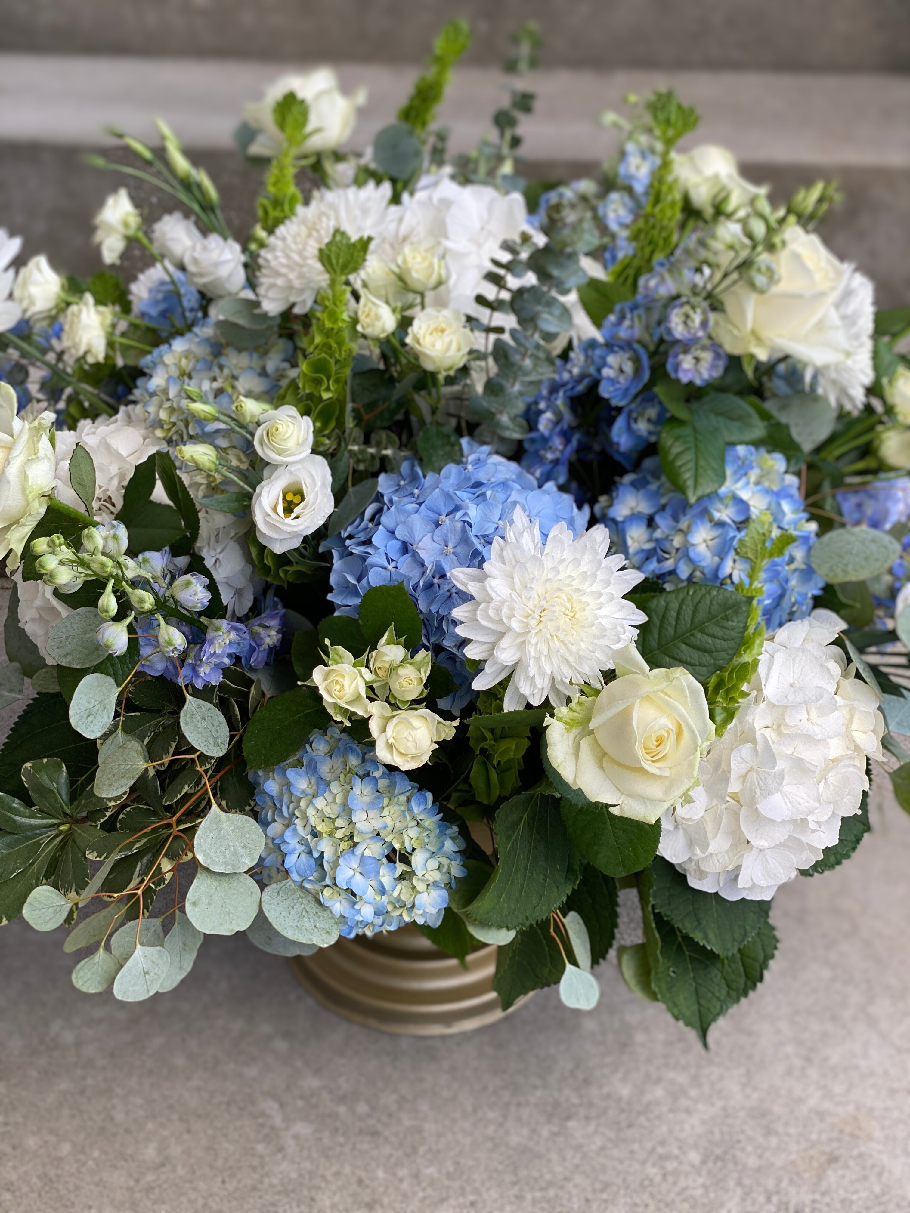 A close up of a large round statement centerpiece created in a gold footed vase and designed with a blue and white colour palette featuring hydrangeas, garden roses, dahlias, delphinium, spray roses, lisianthus, green bells of ireland and accented with pittosporum leaves, and silver dollar and seeded eucalyptus.