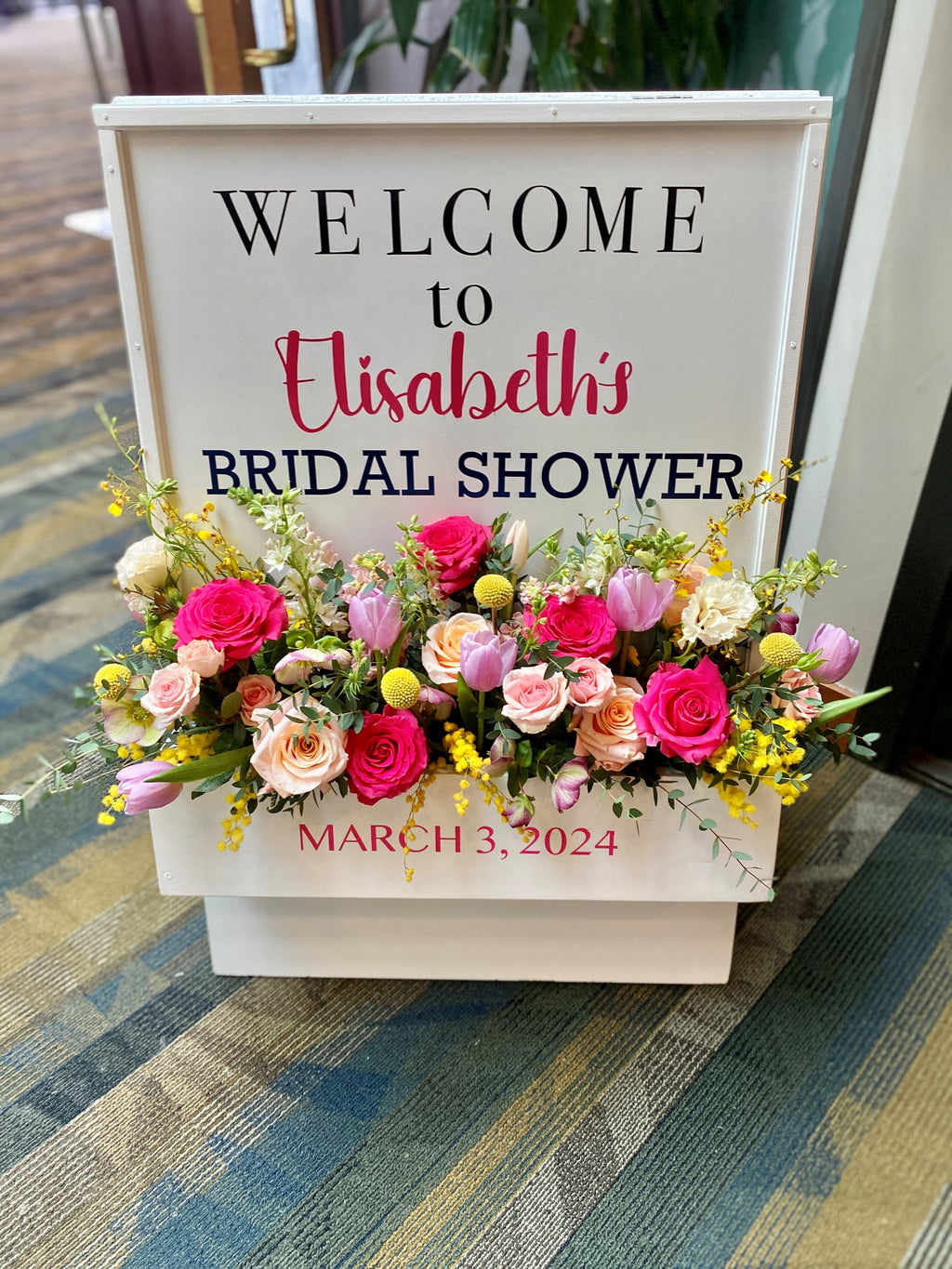 Decorative white wooden  "Welcome" sign floral box, accented with vibrant spring flowers containing fuchsia roses, peach roses, pink tulips, yellow craspedia balls, yellow oncidium orchids, yellow mimosa, white lisanthus and snap dragons, pink hellebores, and eucalyptus varieties displayed at the entrance to a bridal shower.