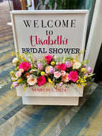 Decorative white wooden  "Welcome" sign floral box, accented with vibrant spring flowers containing fuchsia roses, peach roses, pink tulips, yellow craspedia balls, yellow oncidium orchids, yellow mimosa, white lisanthus and snap dragons, pink hellebores, and eucalyptus varieties displayed at the entrance to a bridal shower.