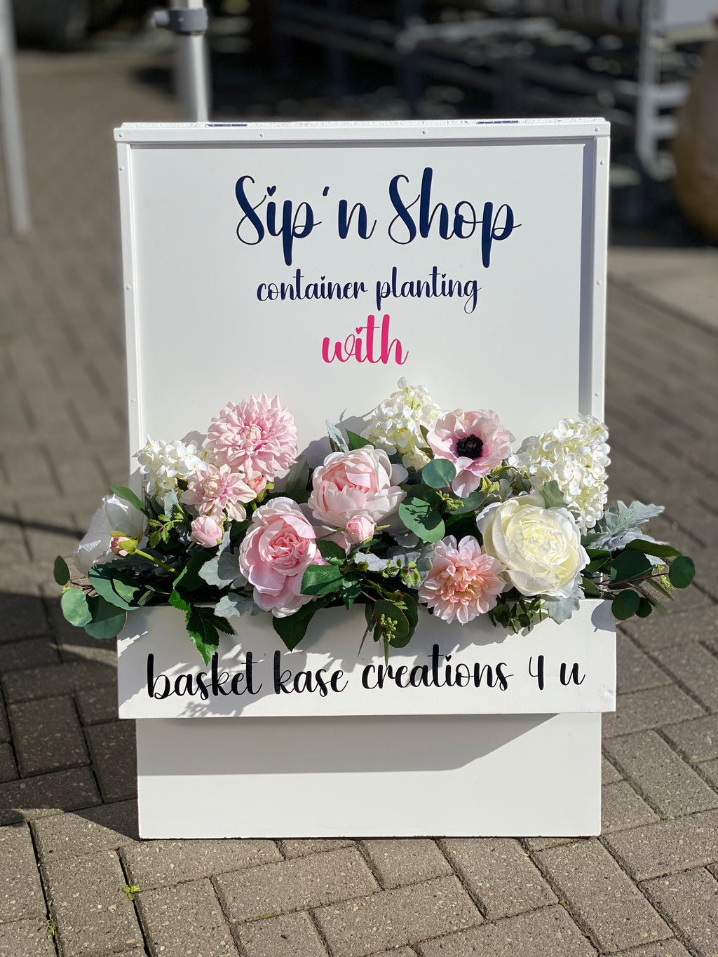 Decorative white wooden sign floral box, accented with white roses, pink peonies, blush anenome, pink dahlias, white hydrangeas, dusty miller leaves and eucalyptus varieties floral arrangement displayed at the entrance to a demonstration event.